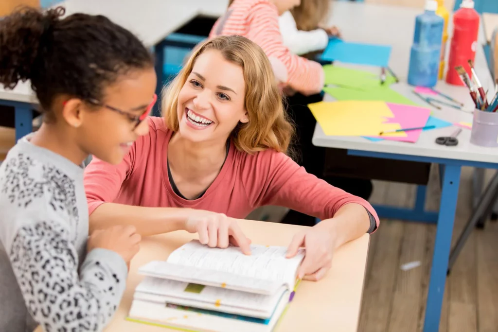 Smiling teacher helping young student with book in classroom setting